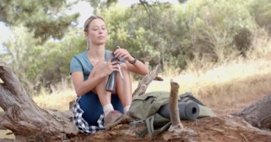 Genç bir kadın devrilmiş bir ağaçta dinleniyor, şişeden su içiyor. Doğa ile çevrili, yürüyüş sırasında bir mola alıyor gibi görünüyor, yanında bir sırt çantası ve minder ile, yavaş çekim.