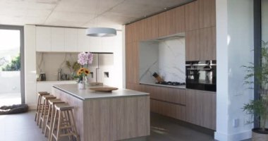 Modern bir mutfakta ahşap tahıl dolabı ve bar tabureleri olan merkezi bir ada bulunur. Güneş ışığı boşluğu dolduruyor, parlak tasarımı vurguluyor ve bir tutam renk katıyor..