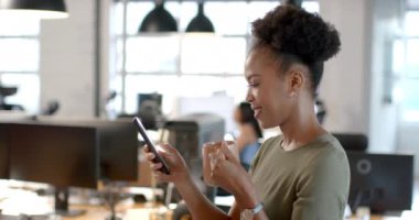 Afrikalı Amerikalı iş kadını telefonunu modern bir ofiste kontrol ediyor. İş arkadaşlarıyla yavaş çekim yaparken kahve molası vermeyi seviyor..