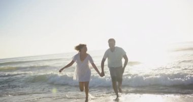 Çift ırklı çift gün batımında plaj boyunca eğlenceli bir koşudan hoşlanır. Beyaz giysileri, akşam ışığının altın rengiyle çelişiyor. Yavaş çekim..