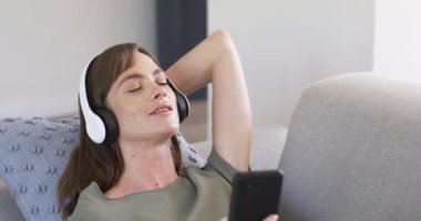 Beyaz kulaklık takmış, gözleri kapalı müzik dinleyen beyaz bir kadın. Kanepede rahat bir şekilde, elinde akıllı bir telefon, yavaş çekimde..
