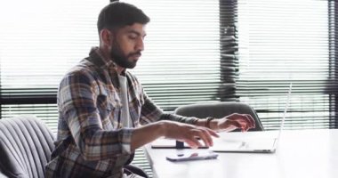 Ekose gömlekli genç Asyalı işadamı ofis ortamında fotokopi alanı ile iş yapmak için akıllı telefon kullanıyor. Cihazına odaklanmış görünüyor. Yakınlarda bir dizüstü bilgisayar var. Bu da işle ilgili bir görev olduğunu gösteriyor..