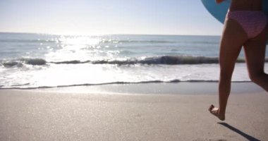 Genç beyaz kadın kolunun altında bir sörf tahtasıyla okyanusa doğru koşuyor. Pembe bir bikini giyiyor, uzun sarı saçları arkasında güneşli kumsal ortamında, ağır çekimde..