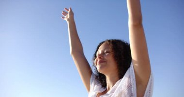 Kıvırcık saçlı melez bir kadın kollarını açık mavi gökyüzüne uzatıyor. Beyaz bir kıyafet giyiyor ve dışarıda özgürlük ve rahatlama hissi yayıyor, ağır çekimde..