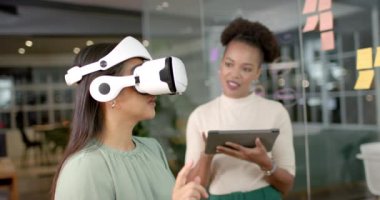 Çift ırklı bir kadın ofis ortamında VR kulaklığı dener, fotokopi alanı ile. Genç Afrikalı Amerikalı kadın asistanlık yapıyor, iş yerinde yenilikçi teknolojinin altını çiziyor, yavaş çekim.
