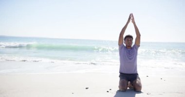 Genç, melez bir adam, fotokopi alanı olan güneşli bir plajda yoga yapıyor. Elleri başının üstünde sıkıca bağlı, ve okyanus dalgaları sakin bir arka plan sağlıyor, yavaş çekim..