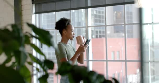 アフリカ系アメリカ人の実業家が窓のそばに立ち コピースペースがある 彼女は明るいオフィスの設定で彼女の携帯電話にテキストしているように見えます スローモーション — ストック動画