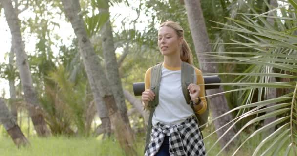若い白人女性は 笑顔で周りを見回して緑豊かな公園をハイキングします 彼女はバックパックとカジュアルな屋外服を着て 自然の中で晴れた日を楽しんで ゆっくりとした動き — ストック動画