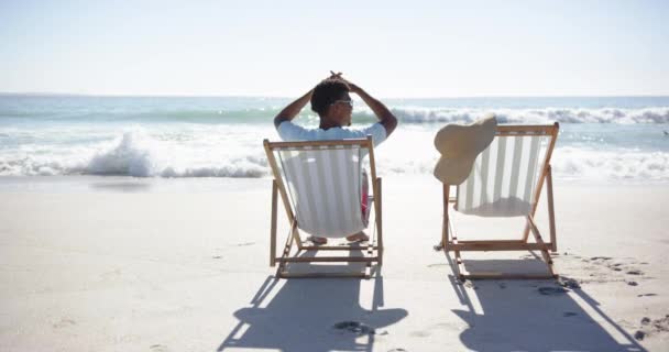 Jovem Biracial Relaxa Uma Cadeira Praia Frente Para Oceano Sugerindo — Vídeo de Stock