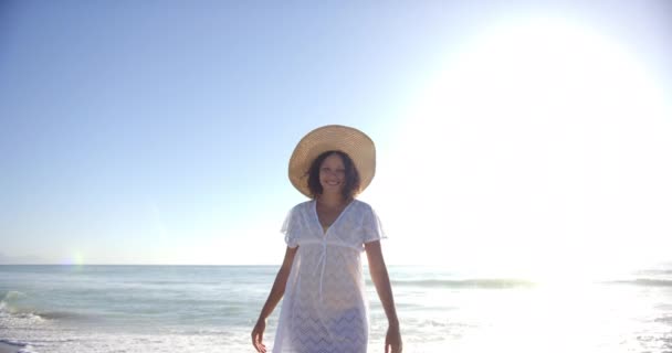 若い女性がビーチに立って 広いレンガの帽子と白いカバーを被っている 彼女の笑顔は 明るい太陽と穏やかな海の波の背景に対して放射する — ストック動画