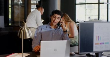 Modern ofiste dizüstü bilgisayarla çalışırken akıllı telefondan konuşan genç bir profesyonel. İş, iletişim, teknoloji, çalışma alanı, çoklu görev, kurumsal