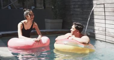 Arka bahçedeki havuzda pembe ve çizgili yüzüklere yaslanan çeşitli gülümseyen çift, su borusuyla. Havuz, eğlence, rahatlama, canlı, açık hava, romantizm, yaşam tarzı