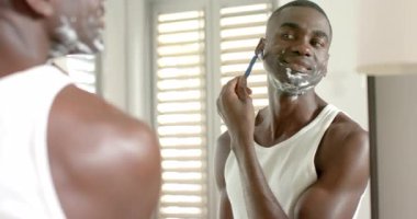 Orta yaşlı Afro-Amerikan erkek tıraş köpüğünü banyoda havluyla, aynayla, güneş ışığıyla siliyor. Cilt bakımı, bakım, yansıma, temizlik, sağlık, iç mekan, doğal ışık.