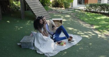 Çeşitli çiftler bahçede pikniğin tadını çıkarıyorlar. Meyve tabağıyla battaniyeye selfie çekiyorlar. Doğa, rahatlık, yaz, rahatlama.