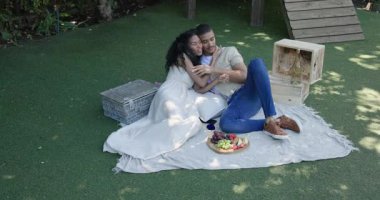 Çeşitli çiftler piknik battaniyesinde dinleniyor, meyvenin tadını çıkarıyor ve bahçede akıllı telefonlara bakıyor. Boş zaman, rahatlama, teknoloji, yaşam tarzı, bağlanma