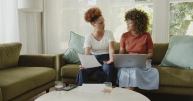 Evde, oturma odasındaki kanepede dizüstü bilgisayar kullanırken aynı cinsiyetten lezbiyen çift belgeleri tartışıyor. LGBTQ, ortaklık, tartışma, teknoloji, ev, iletişim