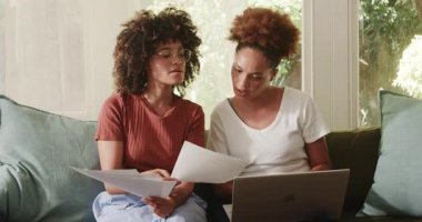 Evrakları gözden geçirip dizüstü bilgisayar kullanarak evdeki kanepede çeşitli lezbiyen çiftler. Uzaktan çalışma, ev ofisi, çoklu görev, teknoloji, gündelik, modern