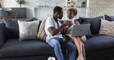 Dizüstü bilgisayara doğru hareket eden, oturma odasındaki kanepede beyaz kupa ve yastıklarla oturan çift. Rahat, modern, yaşam tarzı, iç mekan, rahatlatıcı, dostluk, ev