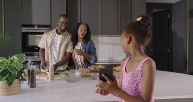 Modern mutfakta ebeveynler birlikte yemek pişirirken akıllı telefona gülümseyen genç bir kız. Çeşitli aile, teknoloji, bağlılık, mutluluk, ebeveynlik, yaşam tarzı