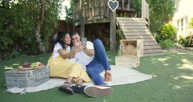 Çeşitli çiftler bahçede piknik battaniyesinde dinleniyor, akıllı telefonlarla neşeli anları paylaşıyorlar. Rahatlama, rahatlama, teknoloji, bağlanma, birliktelik, hafta sonu