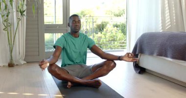 Yatak odasında yoga minderinin üzerinde bambu bitkisiyle bağlı oturan Afrikalı Amerikalı bir adamı meditasyon yaparken. Sağlık, sükunet, farkındalık, iç mekan, dinginlik, rahatlık, rahatlama.