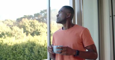 Gülümseyen, orta yaşlı, Afro-Amerikan bir adam, elinde açık mavi kupa, sürgülü veranda kapısında, fotokopi alanında. Rahat, açık hava, boş zaman, doğa, sükunet, yaşam tarzı, modern