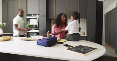 Anne mutfakta öğle yemeği hazırlarken baba okul üniformalı kızı kaldırıyor. Çeşitli aileler, ebeveynlik, çocukluk, yaşam tarzı, birliktelik, sabah rutini