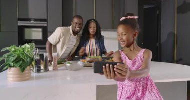 Kızları tabletle fotoğraf çekerken, çeşitli aileler modern mutfakta birlikte yemek yapıyor. Çeşitli aile zamanları, teknoloji, yaşam tarzı, birliktelik, samimiyet, bağlanma