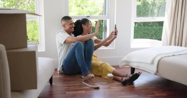 Zeminde oturan çeşitli çiftler yeni evlerinde kutularını yanlarında taşıyarak selfie çekiyorlar. Yer değiştirme, paketleri açma, yeni başlangıçlar, akıllı telefon, yaşam tarzı, mutluluk