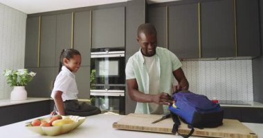 Baba, modern mutfakta kızının sırt çantasını topluyor ve birlikte mutlu bir şekilde gülümsüyor. Çeşitli aileler, bağlılık, ebeveynlik, yaşam tarzı, hazırlık, sabah rutini