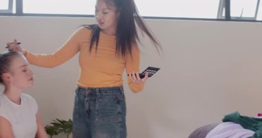 Kadın arkadaşlar makyaj sanatçısını yatakta karşılıyor, göz farı takıyor, kıyafetleri sınıflandırıyor, telefonunu paylaşıyor. Arkadaşlık, yaşam tarzı, gündelik, canlı, sosyal, kendini ifade etme, güzellik