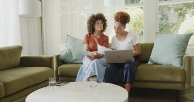 Lezbiyen çift evdeki parlak oturma odasında dizüstü bilgisayarlarıyla belgeleri gözden geçiriyorlar. işbirliği, takım çalışması, uzaktan çalışma, yaşam tarzı, kapalı alan, gündelik