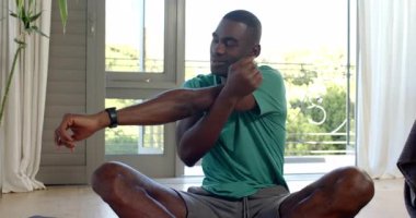 Esneyen Afrikalı Amerikalı bir adam, yoga minderiyle birlikte çalışma alanında bağdaş kurarak yerde oturuyor. Fitness, sağlık, dinginlik, ev spor salonu, rahatlama, farkındalık, modern