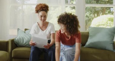 Lezbiyen çift koltukta oturup belgelere bakıyor, gülümsüyor ve evde eğleniyorlar. finans, bütçe, evrak işleri, mutluluk, birliktelik, yaşam tarzı