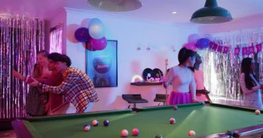 Çeşitli arkadaşlar doğum günü pastasına doğru el kol hareketi yapıyor, selfie çekiyor ve oyun odasında bilardo oynuyor. Kutlama, dostluk, eğlence, coşku, eğlence, sosyalleşme, şenlik