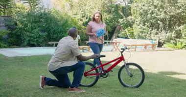 Afrikalı Amerikalı baba oğluna kırmızı bisiklet sürmeyi öğretiyor. Çeşitli anneler arka bahçede miğfer tutuyor. ebeveynlik, öğrenme, çocukluk, aile, güvenlik, destek