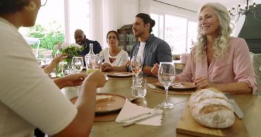 Çeşitli arkadaşlar yemek masasında oturmuş ekmek dilimleyen, tabakları uzatan ve tost yapan kadınları izliyorlar. Samimi, çok kültürlü, neşeli, kırsal, kutlama, doğal ışık, sıcak