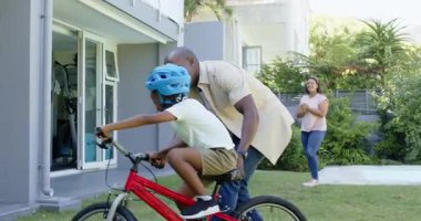 Afrikalı Amerikalı baba oğluna bisiklet sürmeyi öğretiyor, ikisi de gülümsüyor. Ebeveynlik, akıl hocalığı, çocukluk, mutluluk, çeşitli aileler, öğrenme