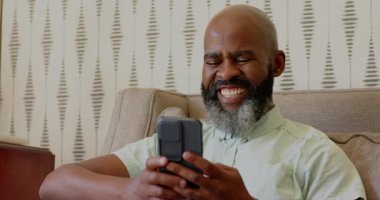 Kıdemli Afrikalı Amerikalı adam oturma odasındaki kanepede akıllı telefon kaydırıyor, gülüyor, suyu yudumluyor. Olgun, rahatlama, yaşam tarzı, rahat, rahat, teknoloji, iç mekan