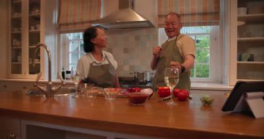 Mutfakta, kıdemli Asyalı çift evde yemek malzemeleri ve tabletlerle dans ediyor. emeklilik, yaşam tarzı, mutluluk, dans, teknoloji, yerli