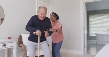 Asyalı bir kadın, yaşlı bir adamın evdeki sandalyesinde rahat oturmasına yardım ediyor. Bakım, yardım, yaşlılar, destek, sağlık, kıdemli bakım