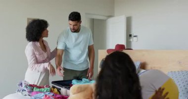 Farklı anneler yataklarına bavul yerleştirirken, kız da evde oyuncak ayıyla izliyor. Seyahat, çeşitli aileler, bavullar, hazırlık, bağ, yolculuk