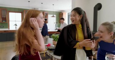 Çeşitli arkadaşlar akıllı telefonun etrafında toplanıyor, mezunlar fotoğraf gösterip mutfakta gülüyor. Kutlama, yoldaşlık, arkadaşlık, modern, sıradan, neşeli, gençlik dolu