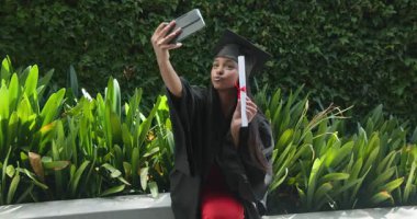 Mezuniyet fotoğrafı için akıllı telefon kaldıran cüppeli Hintli bir kadın. Diploması da bahçe bankında. Kutlama, başarı, yeşillik, neşe, yansıma, açık hava, modern