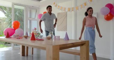 Çeşitli gruplar evde, ahşap masanın yanında renkli pasta ve balonlarla doğum günü partisini kutluyor. Kutlama, doğum günü, şenlik, coşku, atıştırmalıklar, evde, neşe, toplanma, dekorasyon