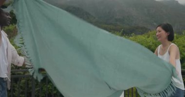 Asyalı kadın ve Afrikalı Amerikalı erkek verandada battaniye sallıyor, yastıkları düzenliyor, piknik hazırlıyorlar. Romantik, boş, samimi, sıradan, açık hava, canlı, neşeli.