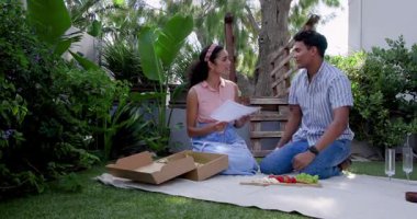 Bahçede yıldönümlerini kutlayan genç bir çift, kumaş üzerinde meyvelerle birlikte kutuyu açıyorlar. Kutlama, romantizm, bahçe, meyve, yıldönümü, açık hava, neşe, çiftlik