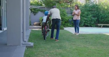 Afrikalı Amerikalı baba oğluna arka bahçede bisiklet sürmeyi öğretiyor, çeşitli anneler izliyor. aile, bağlılık, ebeveynlik, eğitim, çocukluk, açık hava