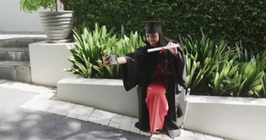 Mezuniyet elbisesi giymiş bir kadın diploma fotoğrafıyla selfie çektikten sonra avluda paylaşmak, fotokopi çekmek. Kutlama, başarı, başarı, mutluluk, açık hava, açık hava, yaşam tarzı