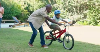 Afrikalı Amerikalı baba çocuğa arka bahçede bisiklet sürmede yardım ediyor, çeşitli anneler tezahürat yapıyor ve alkışlıyor. aile, bağlılık, ebeveynlik, cesaret, çocukluk, açık hava